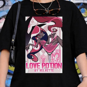 May include: Black t-shirt featuring a graphic of a woman in a martini glass with the text "LOVE POTION BY VELVETTE". The design includes pink and red accents. The shirt is worn by a person.