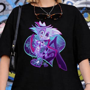 May include: Black t-shirt featuring a cartoon character with a white hat, purple cape, and spiderweb details. The graphic has blue and purple accents. The shirt is worn by a person.