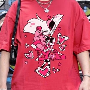 May include: Red t-shirt with a cartoon graphic of two characters embracing, surrounded by hearts. The text "RED COMFORT COLORS" is in the upper left corner. The shirt is worn by a person in front of a graffiti wall.