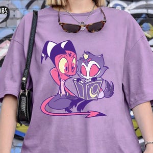 May include: Orchid Comfort Colors t-shirt in a light purple hue. The shirt features a cartoon graphic of two characters, one red and one gray, reading a book. The text "ORCHID COMFORT COLORS" is printed at the top.