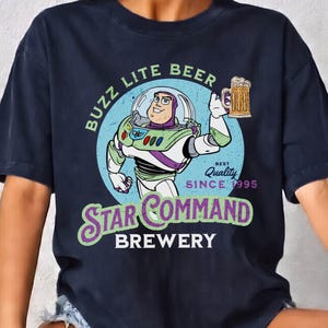 Op de afbeelding: Marineblauw T-shirt met een afbeelding van Buzz Lightyear die een bierpul vasthoudt. Het ontwerp bevat de tekst "Buzz Lite Beer" en "Star Command Brewery" met "Best Quality Since 1995".