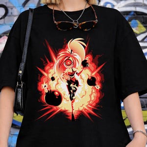 May include: Black t-shirt featuring a vibrant graphic of a character engulfed in an explosion with bombs. The design is in shades of red, orange, and yellow. The shirt is worn by a person in front of a graffiti wall.