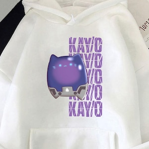 Könnte beinhalten: Weißer Kapuzenpullover mit einer Vordertasche und einer Kapuze mit Kordelzug. Der Pullover zeigt eine lila-blaue Grafik eines katzenartigen Charakters mit dem Wort "KAY/O" vertikal in Lila wiederholt.