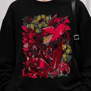 May include: Black sweatshirt featuring a vibrant graphic of a character with red and black accents, surrounded by red roses and yellow flowers. The design has a cartoonish style with geometric shapes and cross details.
