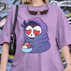May include: Orchid Comfort Colors t-shirt in a light purple hue. The shirt features a cartoon graphic of a character wrapped in a blanket, holding a bowl of ice cream. The text "ORCHID COMFORT COLORS" is printed at the top left.