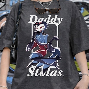 May include: Dark gray t-shirt with the text "Daddy Stolas" and a cartoon graphic of a character in red, blue, and black. The shirt has short sleeves and a relaxed fit. The words "Pepper Comfort Colors" are in the top left corner.