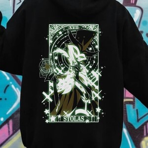 May include: Black hoodie featuring a detailed graphic on the back. The graphic is a stylized illustration with a central figure, surrounded by stars and geometric shapes. The text "XVII" and "STOLAS" are also visible.