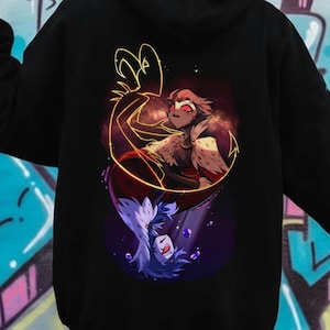 May include: Black hoodie featuring a colorful illustration on the back. The design includes two figures, one with red and brown tones and the other with purple and white tones, surrounded by a golden outline. The hoodie has a hood and long sleeves.