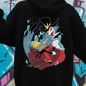 May include: Black hoodie featuring a colorful cartoon illustration on the back. The graphic depicts a character with a crown, holding a book, and wearing a red and black outfit. The design is surrounded by blue and white accents and stars.
