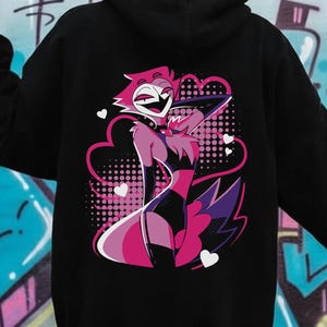 May include: Black hoodie featuring a colorful cartoon character design on the back. The graphic includes pink, purple, and white elements, with a heart-shaped cloud and a dotted background. The hoodie has a hood and long sleeves.