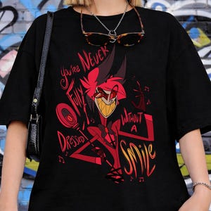 May include: Black t-shirt featuring a red cartoon character with a wide grin and the text "You're Never Fully Dressed Without A Smile." The design includes musical notes and geometric shapes. The shirt is worn by a person.