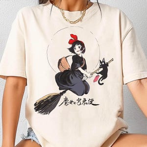 May include: Ivory Comfort Colors t-shirt with a graphic of a young witch and a black cat. The witch is in a black dress, holding a broom. Japanese text is below the graphic. The shirt has short sleeves and is a neutral colour.