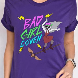May include: A purple t-shirt with the text "BAD GIRL COVEN" in pink and green, with a cartoon illustration of a character riding a broom. The shirt also has the text "TEAM PURPLE BELLA CANVAS".