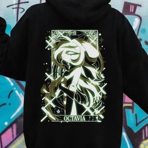 May include: Black hoodie featuring a detailed graphic on the back. The graphic is a stylized illustration with white and olive green accents, and the word "OCTAVIA" is printed at the bottom. The background is a colorful graffiti wall.