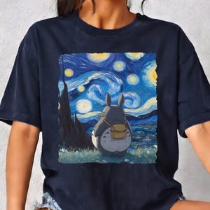 May include: Navy blue t-shirt featuring a graphic of Totoro from My Neighbor Totoro, set against a backdrop inspired by Van Gogh's "Starry Night." The image includes a gray Totoro with a backpack, and the text "NAVY" is in the upper left corner.