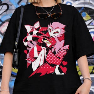 May include: Black t-shirt featuring a graphic of two cartoon characters in red, pink, and white. The design includes hearts and a bow. The shirt is a casual, oversized style.