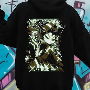 May include: Black hoodie with a graphic design on the back. The design features a stylized character with horns and the word "Husk" at the bottom. The hoodie is worn in front of a graffiti wall.