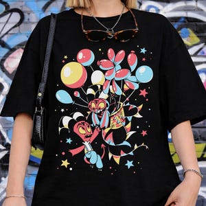 May include: Black t-shirt featuring a colorful cartoon graphic of two characters with balloons, stars, and confetti. The graphic includes red, yellow, blue, and pink elements. The shirt has short sleeves and a relaxed fit.