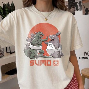 Könnte beinhalten: Ein elfenbeinfarbenes T-Shirt mit einer Grafik eines grünen Monsters und einer grauen Kreatur, die weiße Sumogürtel tragen, vor einem großen orangefarbenen Kreis. Der Text "SUMO" ist in Orange unter den Figuren gedruckt, zusammen mit einem kleinen quadratischen Symbol.