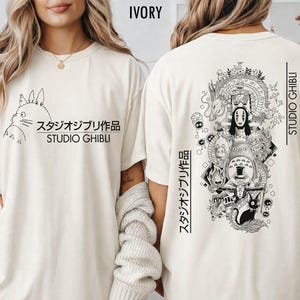 以下が含まれることがあります： アイボリー色のTシャツ。前面には、漫画のキャラクターの白黒イラストと「スタジオジブリ作品 STUDIO GHIBLI」の文字がプリントされています。背面には、キャラクターと「STUDIO GHIBLI」の文字が描かれた詳細なグラフィックがあります。