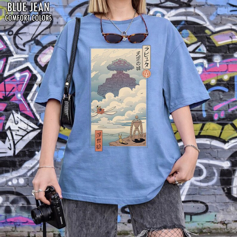 May include: A pale blue t-shirt featuring a Japanese anime-style illustration of a floating castle, a robot, and other figures. The text "BLUE JEAN" and "COMFORT COLORS" is printed at the top of the shirt.