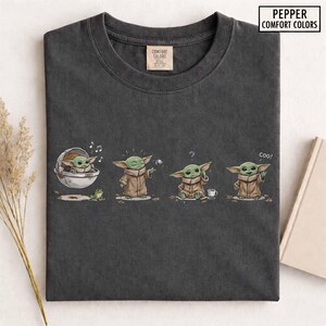 May include: Dark gray t-shirt featuring a cartoon character in various poses. The character is green with large ears and wears a brown cloak. The shirt has the text "Pepper Comfort Colors" in a white rectangle.