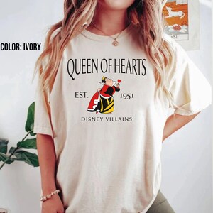 May include: Ivory t-shirt with the text "QUEEN OF HEARTS" and an illustration of the Queen of Hearts from Disney Villains. The graphic includes "EST. 1951" and "DISNEY VILLAINS". The shirt is a light cream colour.