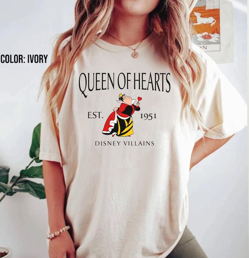 May include: Ivory t-shirt with the text "QUEEN OF HEARTS" and an illustration of the Queen of Hearts from Disney Villains. The graphic includes "EST. 1951" and "DISNEY VILLAINS". The shirt is a light cream colour.