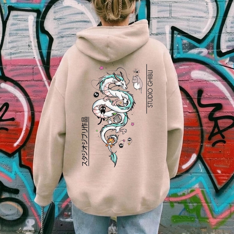 May include: Beige hoodie featuring a detailed illustration of a dragon with turquoise accents, accompanied by Studio Ghibli characters. The back of the hoodie also has Japanese text and the words "STUDIO GHIBLI" in vertical text.