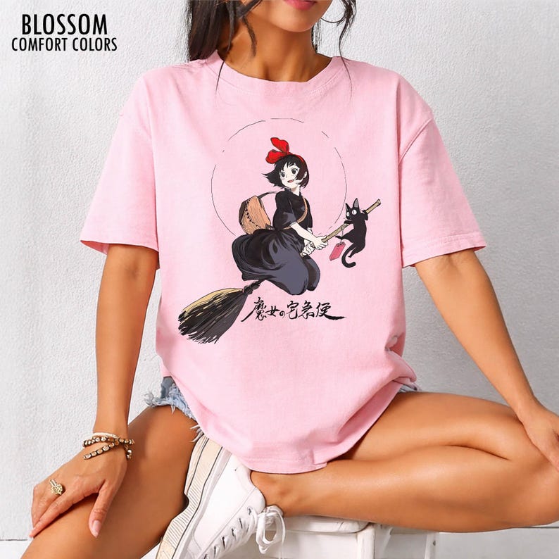 May include: Pink t-shirt with a graphic of a young witch on a broom with a black cat. The witch is wearing a black dress and a red bow. The text "BLOSSOM COMFORT COLORS" is at the top left.