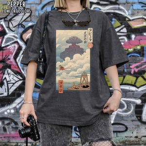 May include: Dark grey t-shirt with a Japanese-style illustration of a floating castle, a robot, and a flying machine. The graphic includes Japanese text. The shirt has short sleeves and a loose fit. The t-shirt is a comfort colors style.