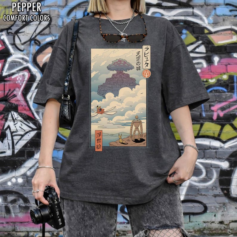May include: Dark grey t-shirt with a Japanese-style illustration of a floating castle, a robot, and a flying machine. The graphic includes Japanese text. The shirt has short sleeves and a loose fit. The t-shirt is a comfort colors style.