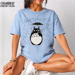 May include: Light blue t-shirt with a graphic of a black and white cartoon character holding an umbrella in the rain. The shirt has the words "CHAMBRAY COMFORT COLORS" printed at the top left.