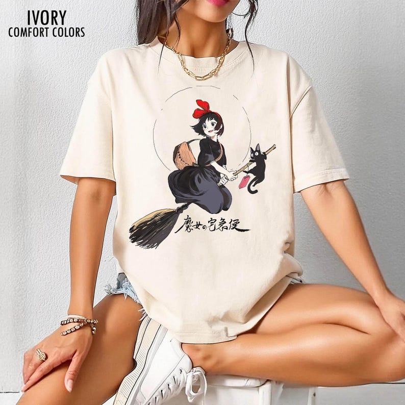 May include: Ivory-colored Comfort Colors t-shirt featuring a graphic of a young witch with a black cat. The witch is wearing a black dress and holding a broom. Japanese text is below the graphic. The shirt has short sleeves.