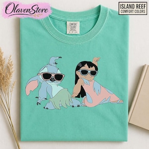 Op de afbeelding: Een turquoise Comfort Colors t-shirt met een afbeelding van twee stripfiguren met een zonnebril. E&eacute;n is blauw met roze oren, de ander is een meisje in een roze jurk. Het shirt heeft de tekst "ISLAND REEF COMFORT COLORS".