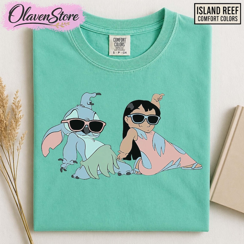 Op de afbeelding: Een turquoise Comfort Colors t-shirt met een afbeelding van twee stripfiguren met een zonnebril. E&eacute;n is blauw met roze oren, de ander is een meisje in een roze jurk. Het shirt heeft de tekst "ISLAND REEF COMFORT COLORS".