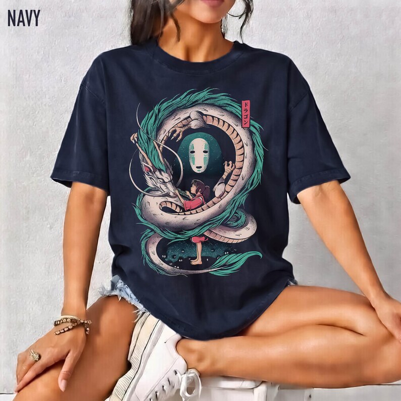 May include: Navy blue t-shirt featuring a detailed illustration of a dragon, a girl, and a masked figure. The design incorporates shades of white, green, and pink. The word "NAVY" is printed at the top left.
