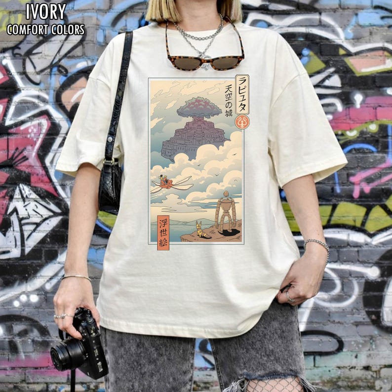 May include: Ivory Comfort Colors t-shirt with a Japanese anime-style illustration. The design shows a floating castle, a robot, and a flying machine. The shirt has short sleeves and a relaxed fit.