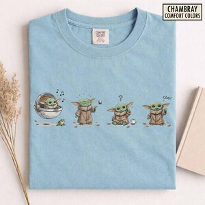 May include: Light blue t-shirt featuring a cartoon character in various poses. The graphic includes the character in a pod, playing with a ball, looking confused, and saying "coo!". The shirt has the text "CHAMBRAY COMFORT COLORS".