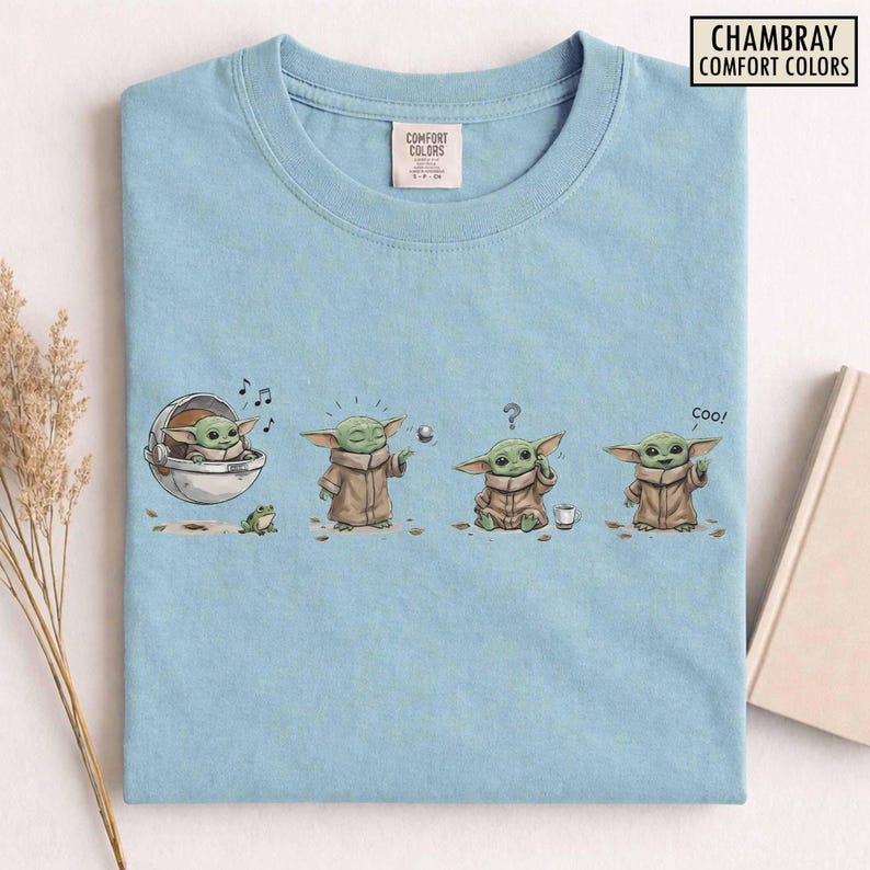 May include: Light blue t-shirt featuring a cartoon character in various poses. The graphic includes the character in a pod, playing with a ball, looking confused, and saying "coo!". The shirt has the text "CHAMBRAY COMFORT COLORS".