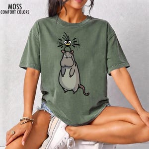 May include: Moss green t-shirt featuring a cartoon hippo and bird design. The hippo is grey with a long tail, and the bird is black with large eyes. The text "MOSS COMFORT COLORS" is printed at the top left.