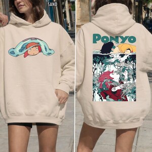 May include: Sand-colored hoodie featuring a front graphic of a cartoon character under a blue blanket. The back of the hoodie displays the word "PONYO" in teal, with a colorful illustration of characters from the animated film.