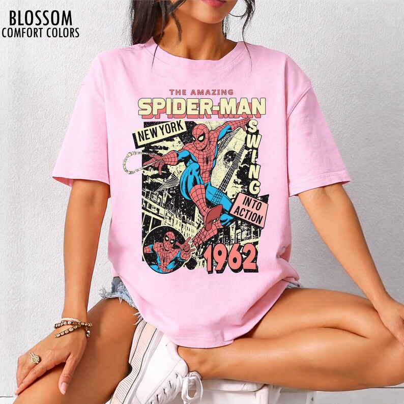 K&ouml;nnte beinhalten: Hellrosa T-Shirt mit einem Vintage-Spider-Man-Design. Der Aufdruck enth&auml;lt die Schriftz&uuml;ge "THE AMAZING SPIDER-MAN", "NEW YORK" und das Jahr "1962". Die Illustration zeigt Spider-Man in Aktion, der durch eine Stadt schwingt.