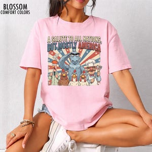 May include: Pink t-shirt with a graphic featuring cartoon characters saluting, with text reading "A Salute to All Nations, But Mostly America." The design includes an eagle, chickens, and other characters in a patriotic theme. The shirt has short sleeves.