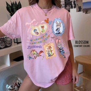 May include: A light pink short-sleeve t-shirt featuring a Zootopia graphic print. The design includes illustrations of Judy Hopps and Nick Wilde, heart-shaped frames, a banner with the text Welcome to Zootopia, and small star accents.