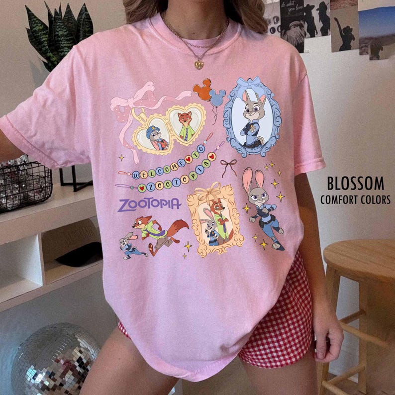 May include: A light pink short-sleeve t-shirt featuring a Zootopia graphic print. The design includes illustrations of Judy Hopps and Nick Wilde, heart-shaped frames, a banner with the text Welcome to Zootopia, and small star accents.