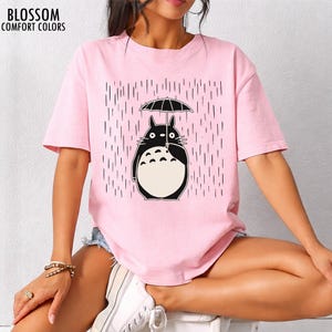 May include: A light pink t-shirt with a black and white graphic of a cartoon character holding an umbrella in the rain. The text "BLOSSOM COMFORT COLORS" is printed at the top left.