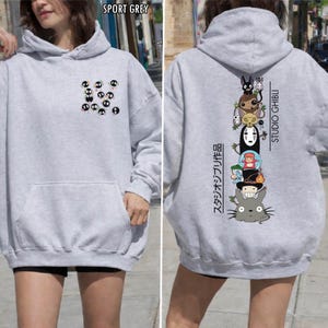 May include: Sport grey hoodie featuring a front pocket and a hood. The front displays a pattern of black and white cartoon characters. The back showcases a vertical stack of Studio Ghibli characters with Japanese text.