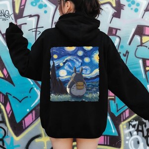 May include: Black hoodie featuring a colorful graphic of Totoro from My Neighbor Totoro, set against a starry night sky reminiscent of Van Gogh's style. The design includes a gray Totoro with a backpack, standing in a field.