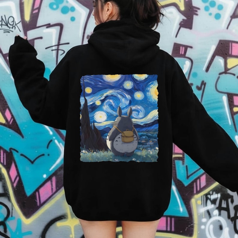 May include: Black hoodie featuring a colorful graphic of Totoro from My Neighbor Totoro, set against a starry night sky reminiscent of Van Gogh's style. The design includes a gray Totoro with a backpack, standing in a field.